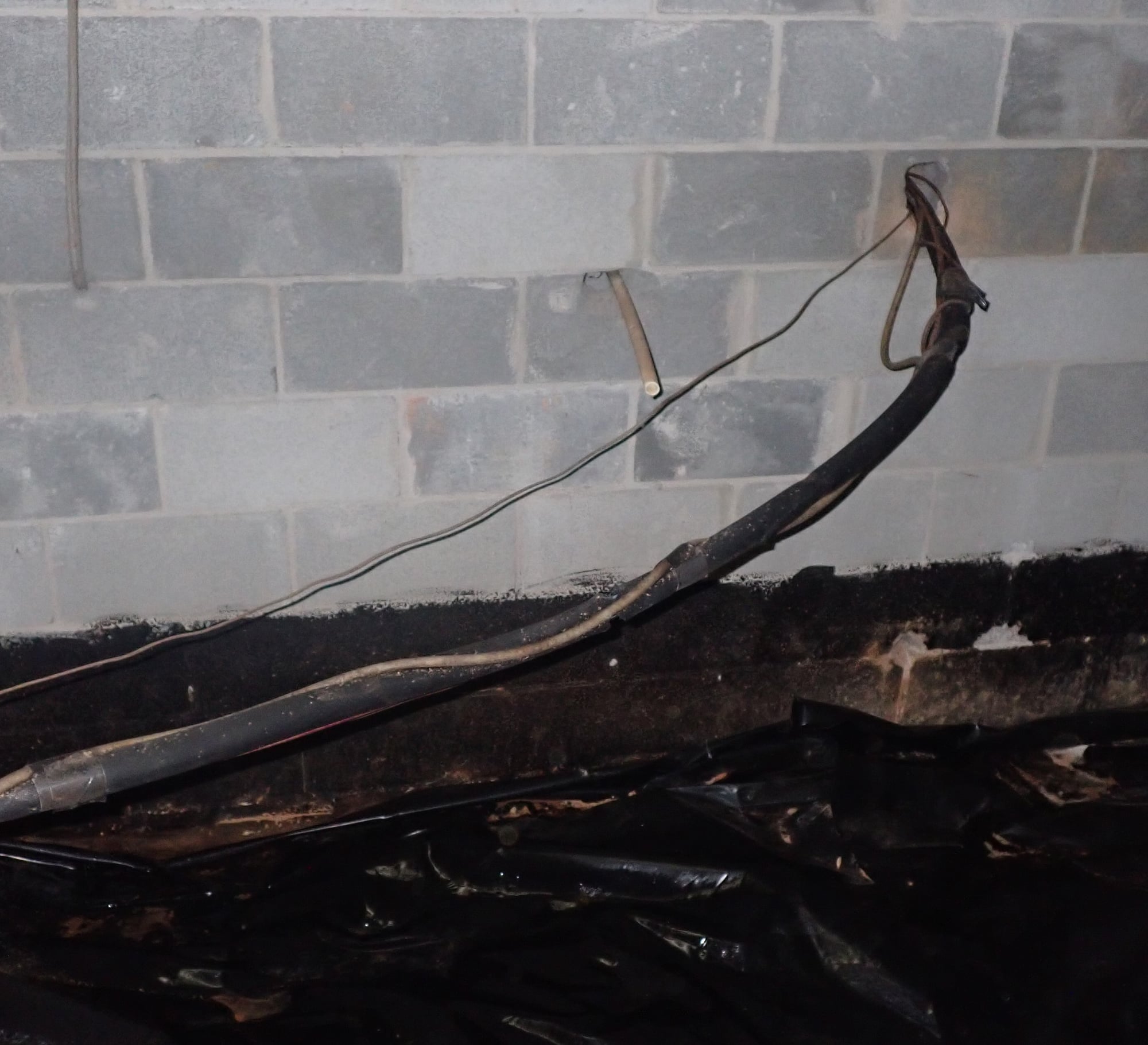Crawlspace foundation wall with a short tan discharge pipe stub protruding and ending in open air above the vapor barrier