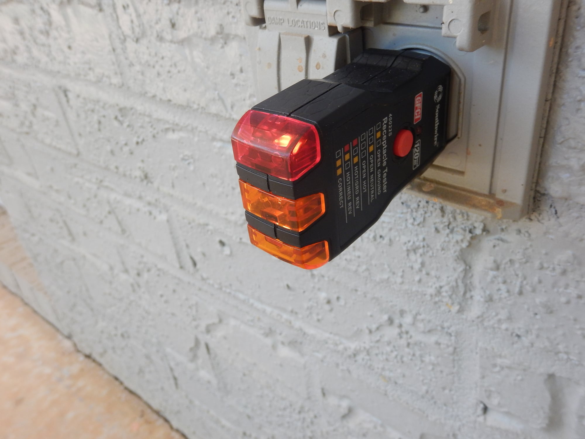 A plug-in receptacle tester in an exterior GFCI outlet showing two yellow lamps lit for hot/neutral reverse, with the weatherproof cover open on a painted block wall