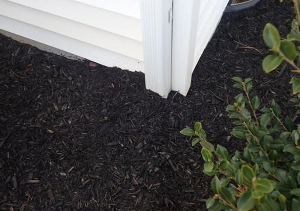 White vinyl siding and downspout at a house corner where dark mulch is piled up against and over the bottom of the siding
